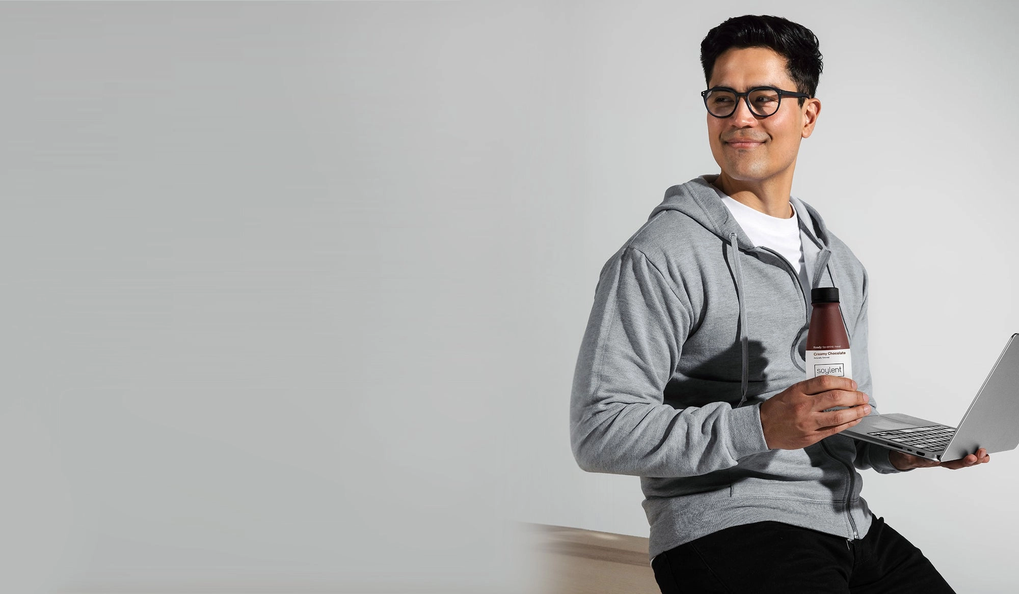 A man in a gray hoodie holding a bottle of Soylent and a laptop