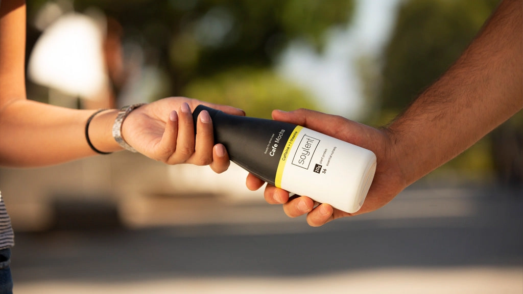 A woman giving a bottle of Soylent to a student in need